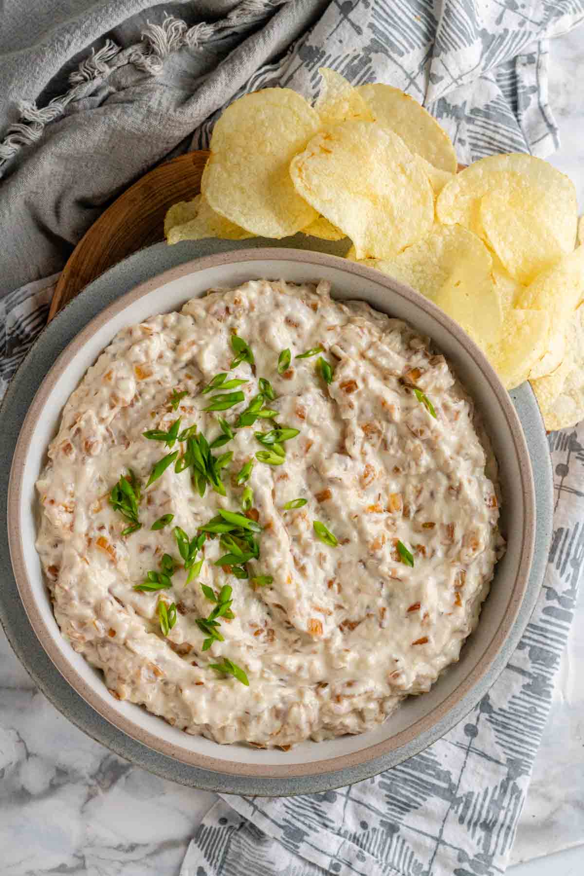 A yogurt dip in a bowl with onions in it and green onions on top beside potato chips.