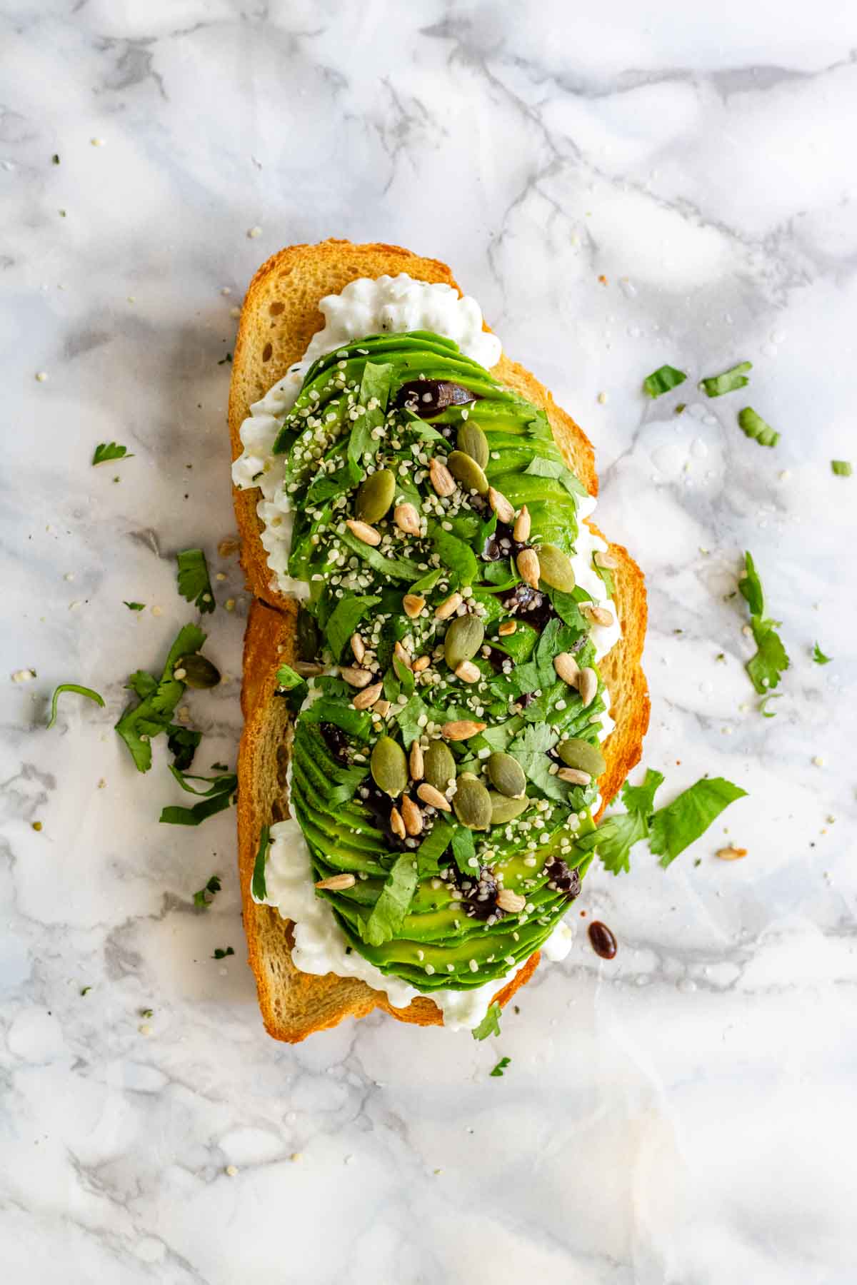 A slice of toast with cottage cheese, sliced avocado, seeds, and cilantro on a white marble background.