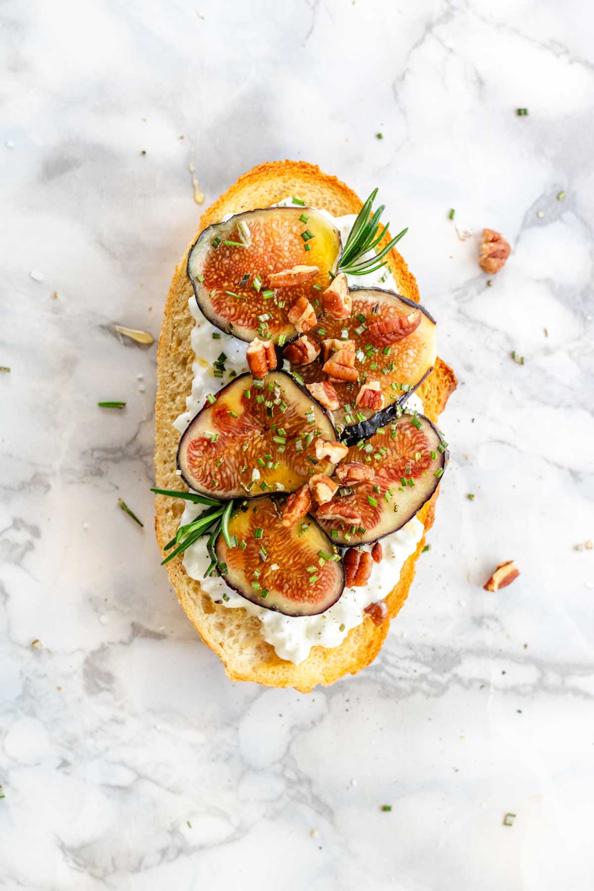 A slice of toast with cottage cheese, figs, rosemary, pecans, and honey on a white marble background.