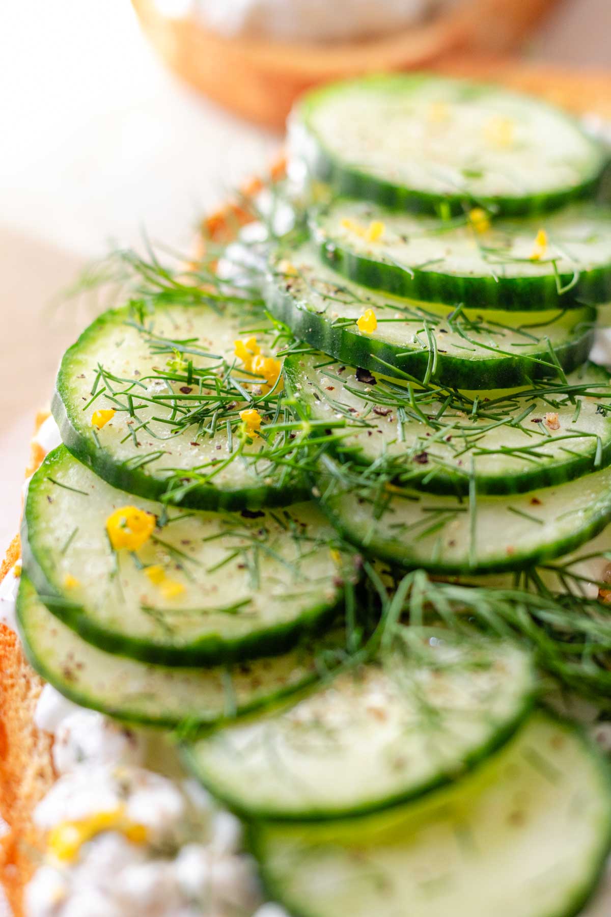 A slice of toast with cottage cheese, sliced cucumbers, lemon zest, and dill on a white marble background.
