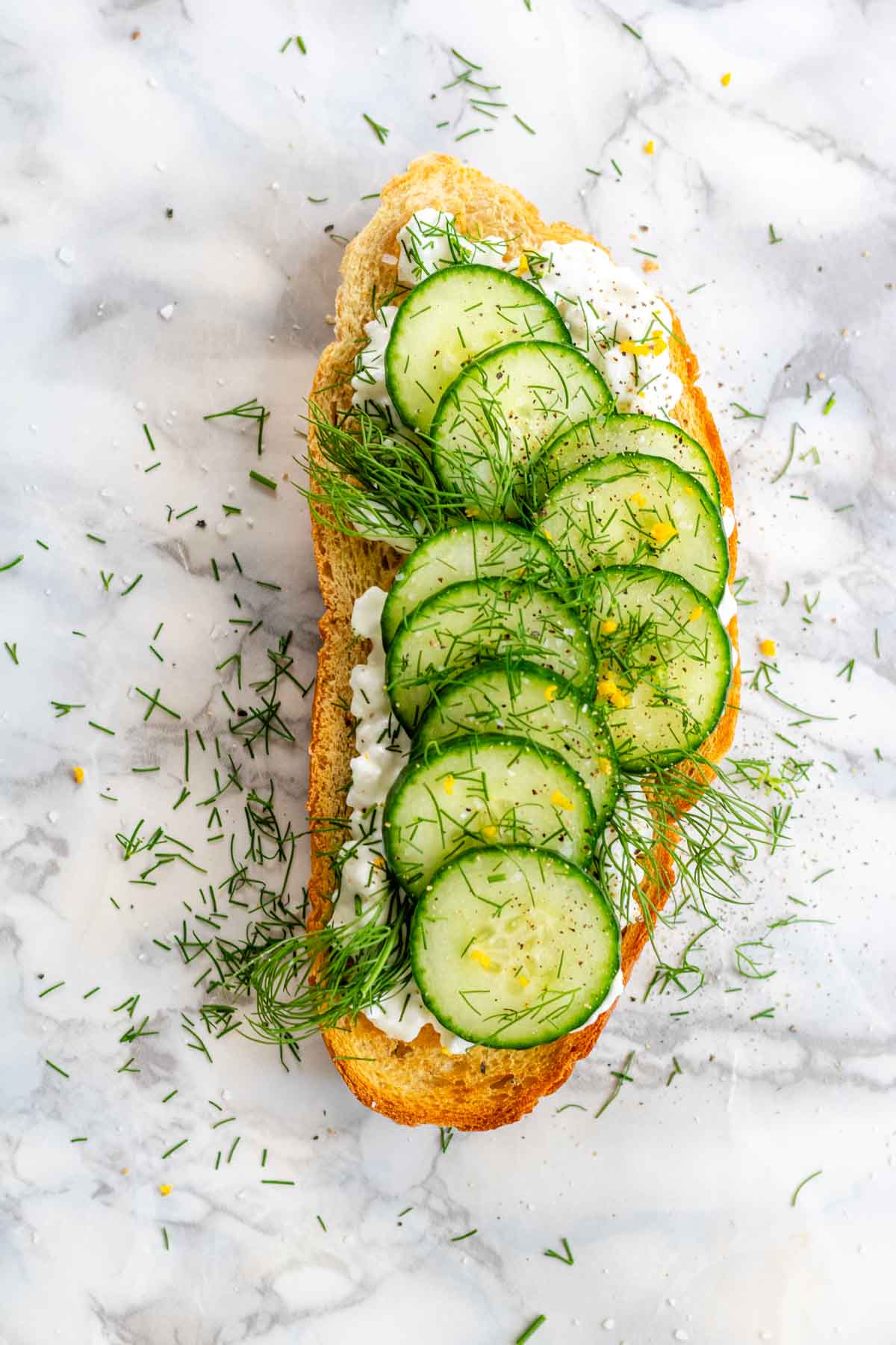 A slice of toast with cottage cheese, sliced cucumbers, lemon zest, and dill on a white marble background.