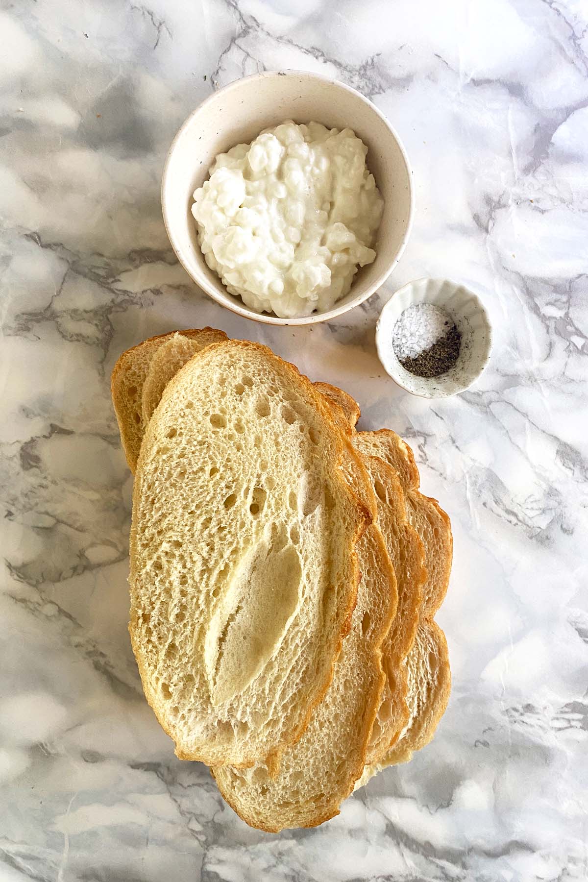 A slice of toast, a bowl of cottage cheese, salt, and pepper, on a white marble background.