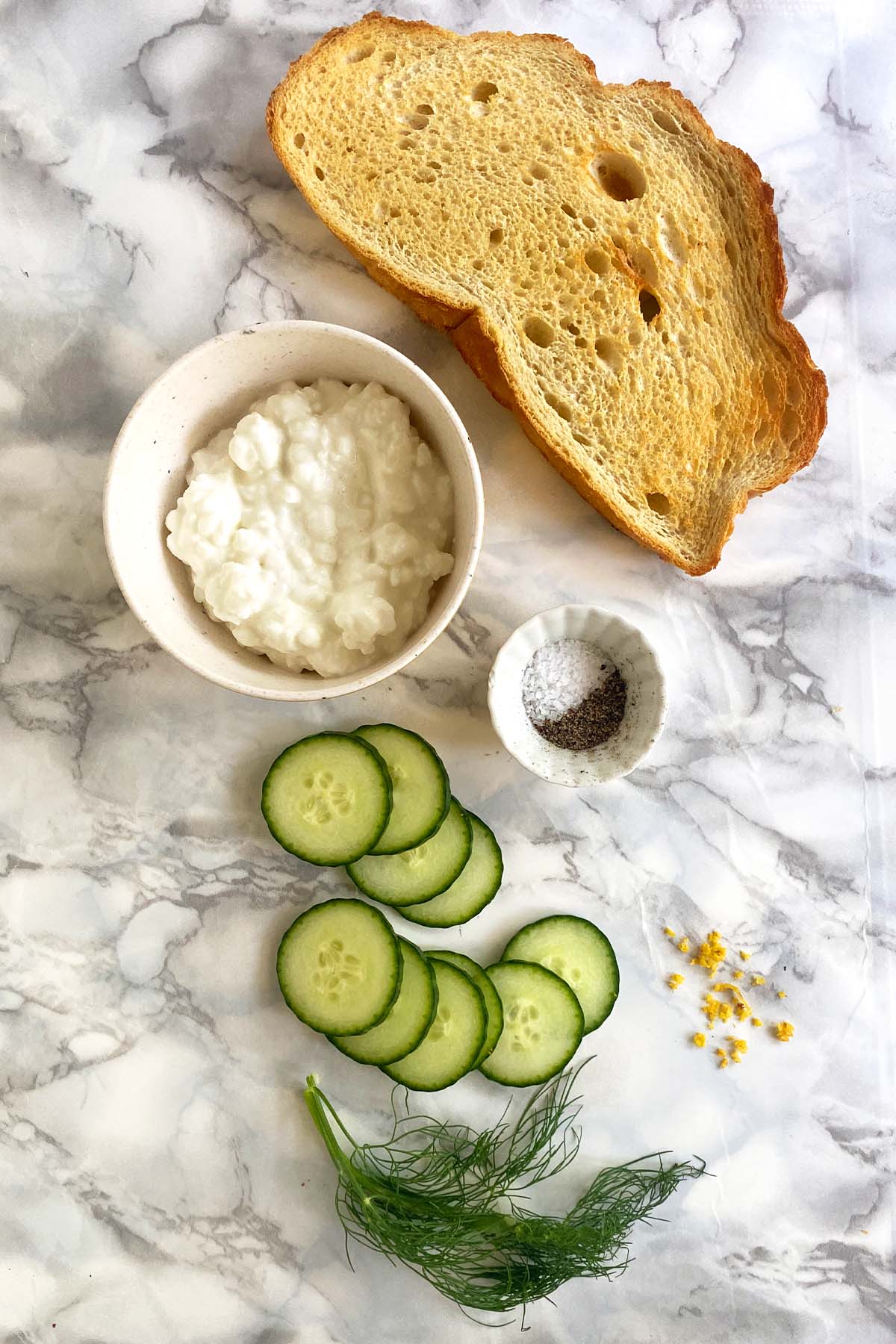 A slice of toast, a bowl of cottage cheese, salt and pepper, sliced cucumber, dill, and lemon zest on a white marble background.