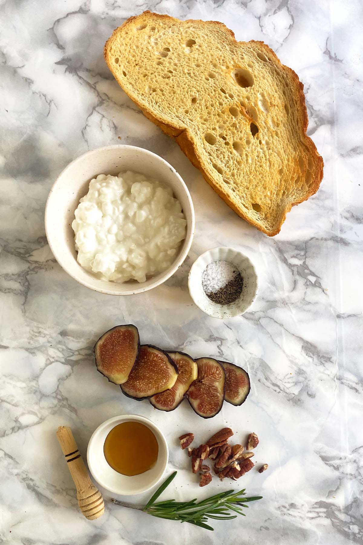 A slice of toast, a bowl of cottage cheese, salt and pepper, sliced fig, rosemary, honey, and pecans on a white marble background.