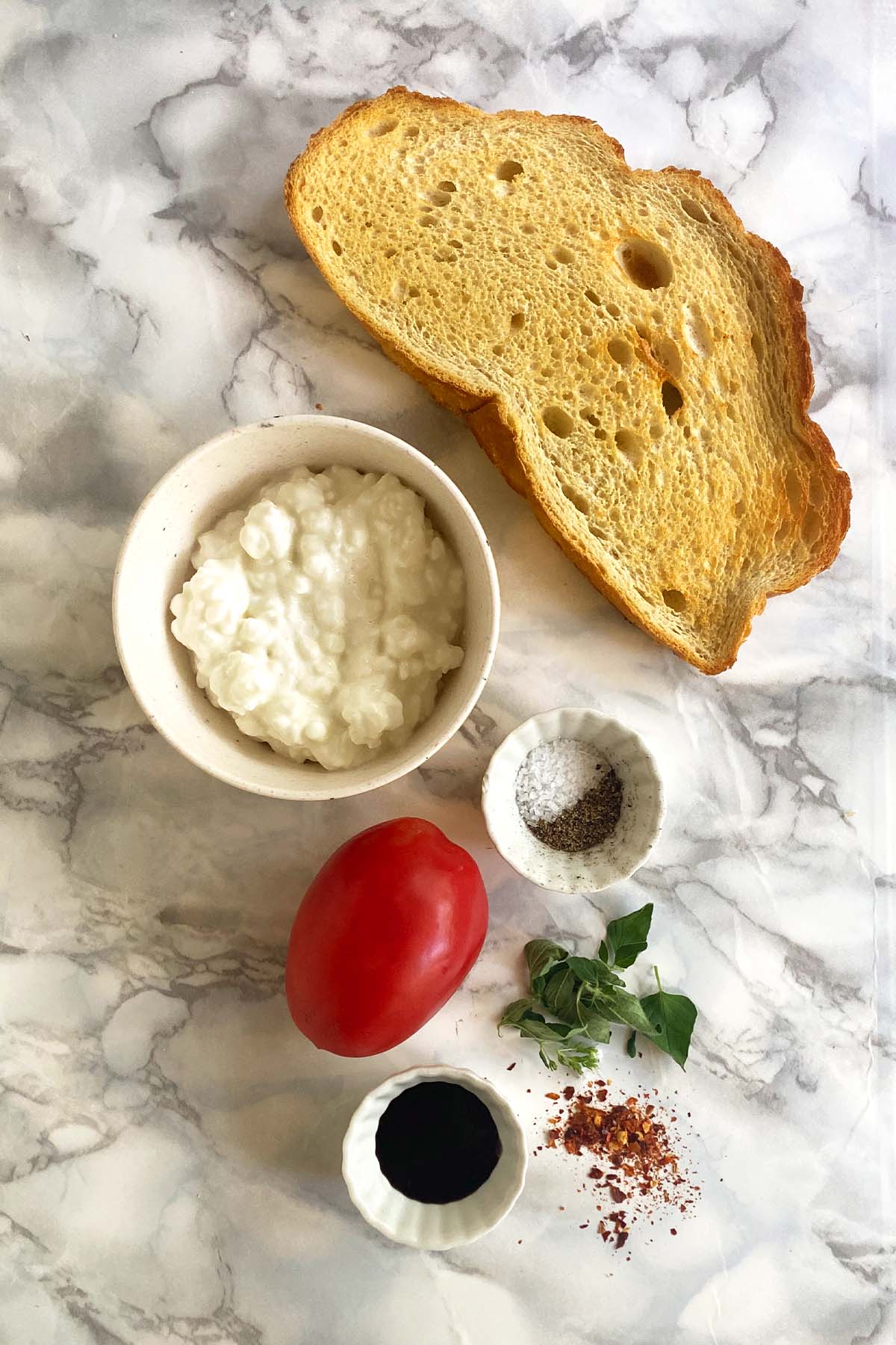 A slice of toast, a bowl of cottage cheese, salt and pepper, a tomato, oregano, red pepper flakes, and balsamic glaze on a white marble background.
