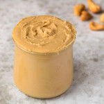 Cashew butter in a clear jar with cashews on the surface around the jar.