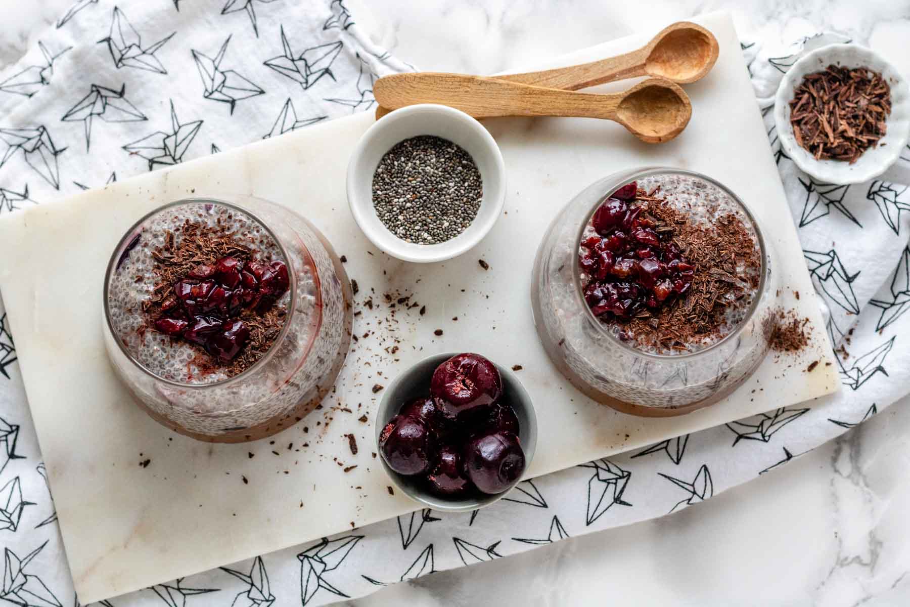 Two glasses with chocolate cherry chia pudding and small bowls of garnishes surrounding them.
