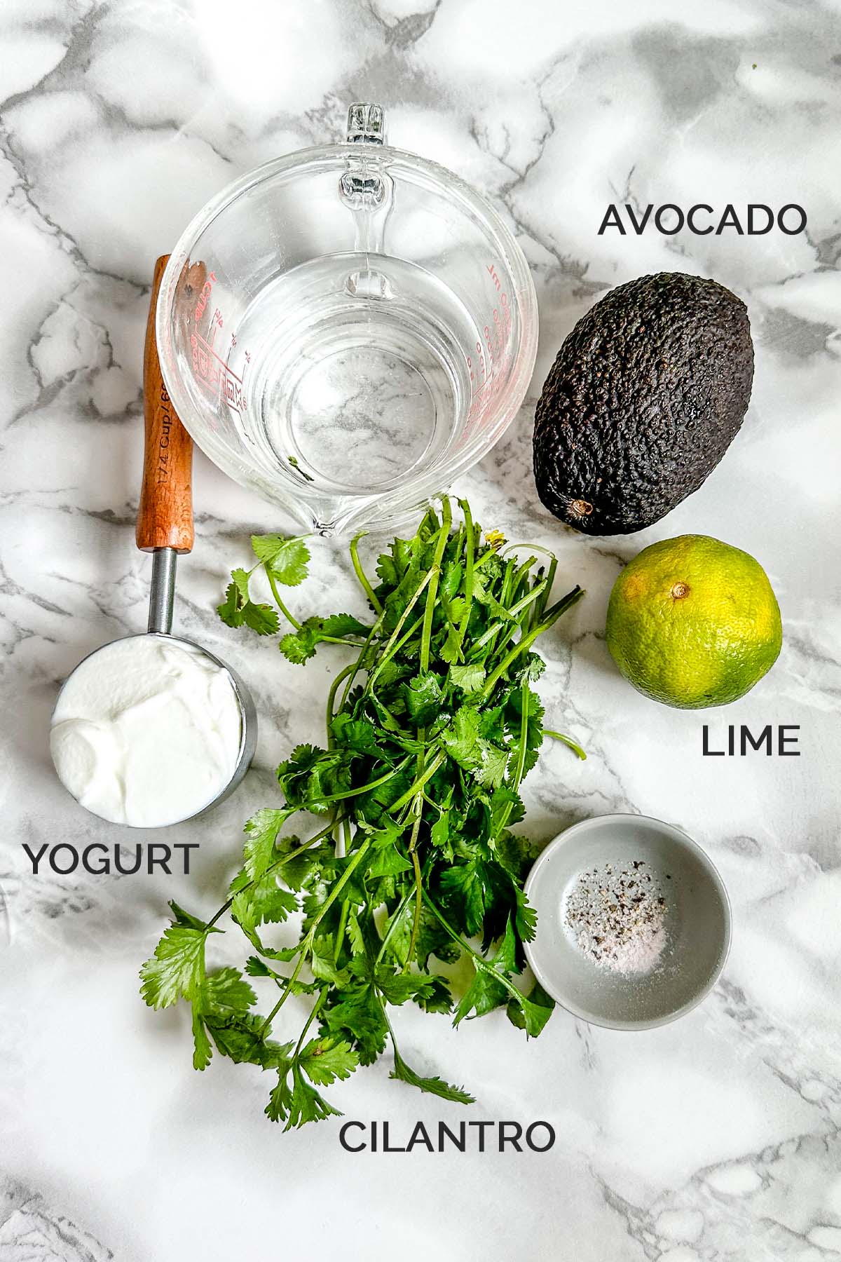 Avocado, lime, cilantro, a cup of yogurt, salt and pepper, and water on a white marble counter.