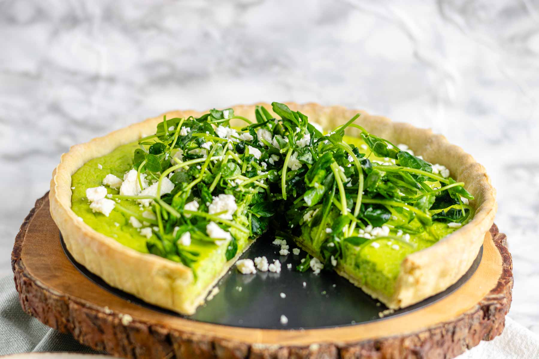 Quiche on a platter with a green filling topped with pea shoots and feta cheese.