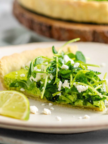 Quiche on a plate with a green filling topped with pea shoots and feta cheese.