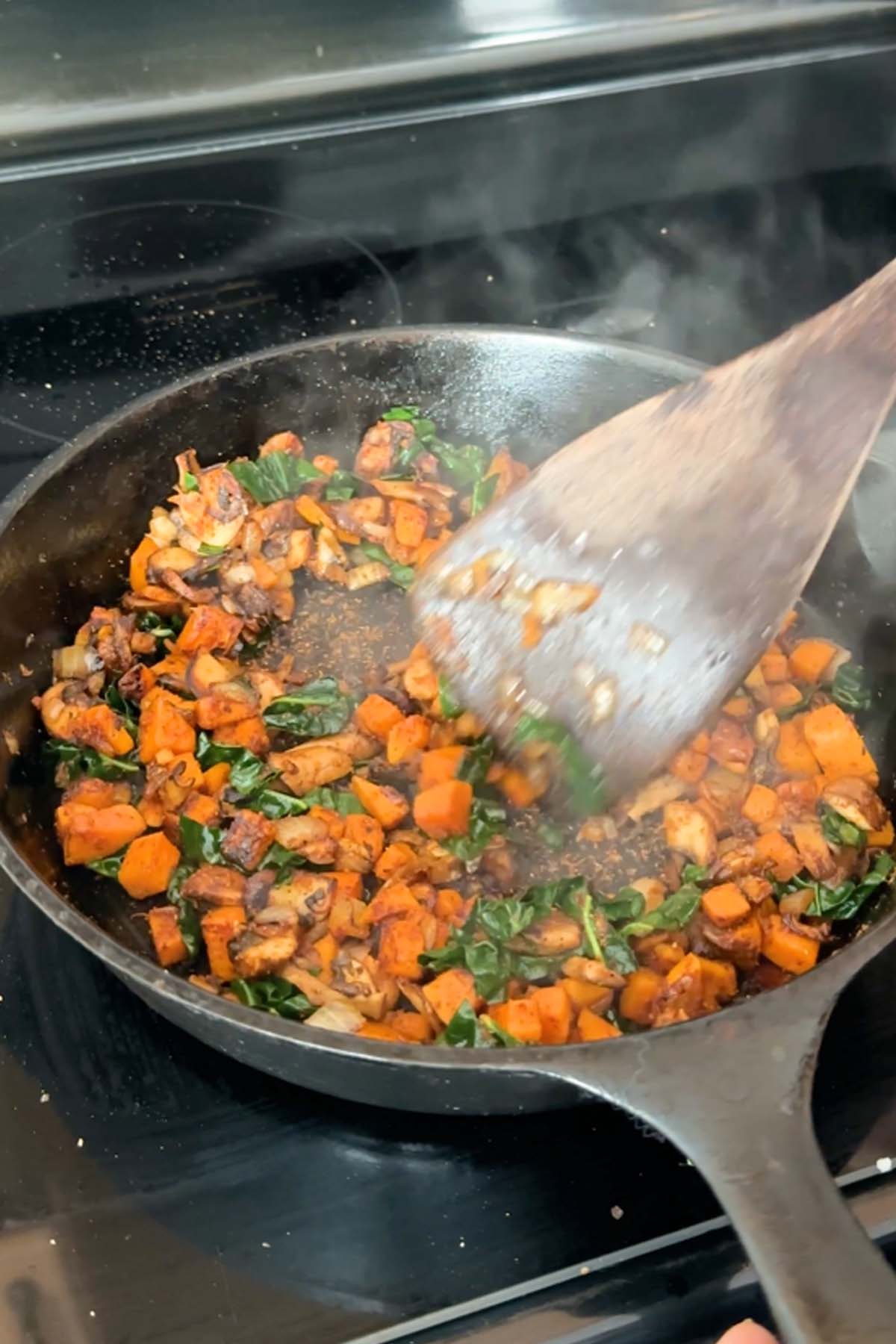 A wooden spoon stirs cubed sweet potato, kale, and sliced mushrooms in a cast iron skillet.
