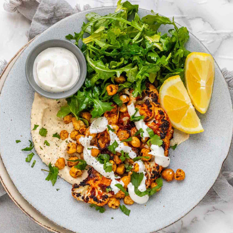 A plate with hummus, cauliflower, chickpeas, arugula, a lemon wedge, and a bowl of white dip.