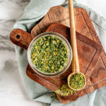 A seasoning mix of yellow and green in a jar with a wooden spoon full of seasoning mix beside it.