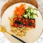 A plate with bulgur topped with a salmon fillet and carrots and cucumber on the side with a fork.