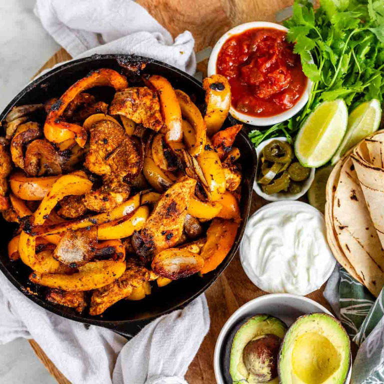 A skillet with grilled strips of chicken and peppers beside bowls of sauce and toppings.