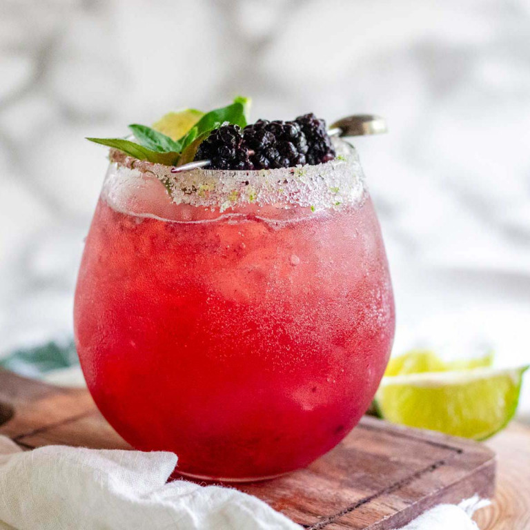 A reddish cocktail garnished with blackberries on a pick, fresh herbs and a lime wedge.