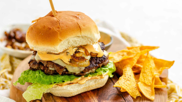 A burger on a bun with lettuce, sauce, cheese, and onions, beside tortilla chips.