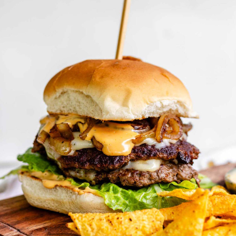 A burger topped with fried onions, sauce, cheese, and lettuce beside tortilla chips.
