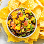 Salad with black beans, mango, tomato, and cilantro in a bowl surrounded by tortilla chips.