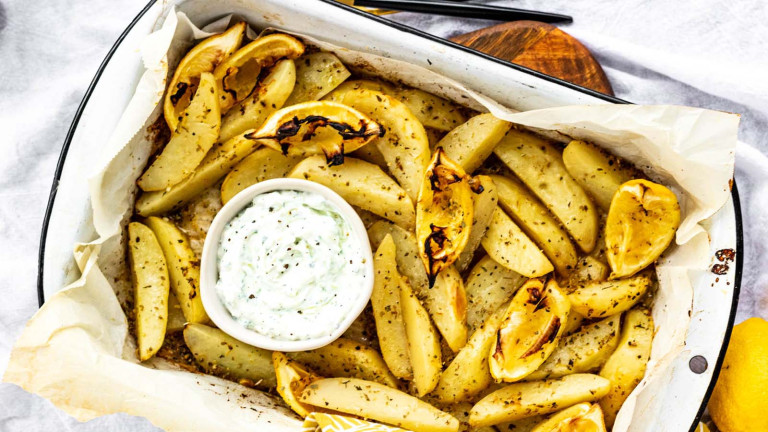 Potatoes wedges in a baking dish with lemon slices and a bowl of dip.