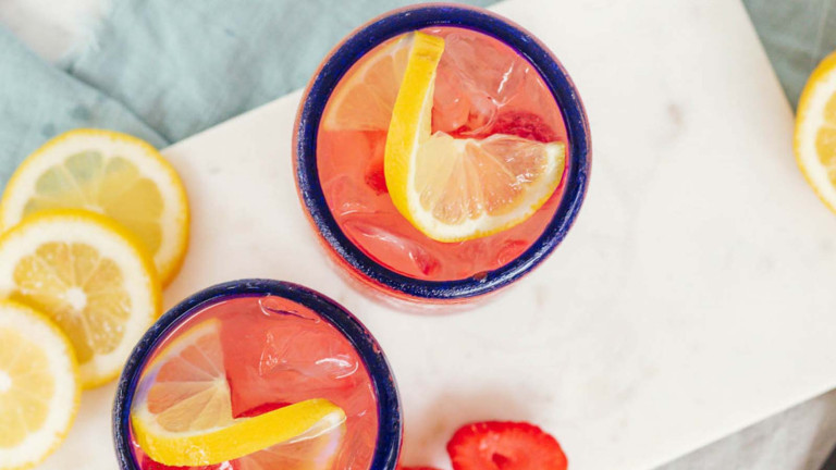 A pink drink in a glass with a blue rim, garnished with lemon slices and strawberries.