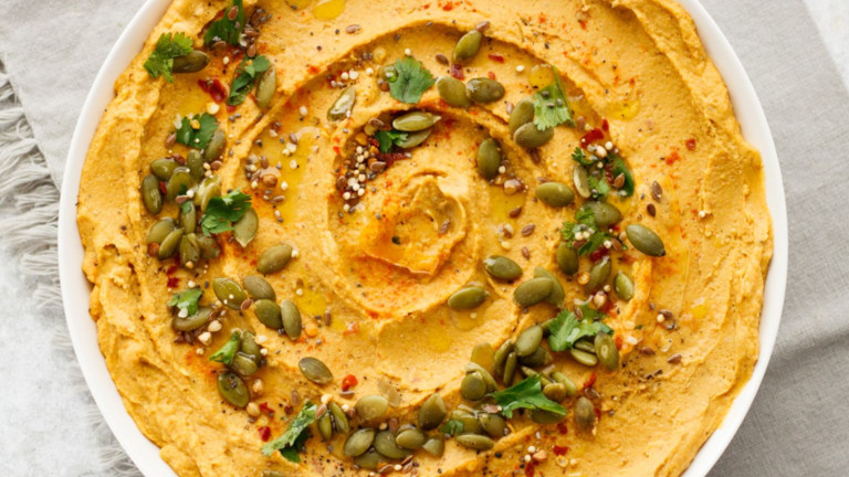 Orange hummus in a bowl with a swirl of seeds on top.