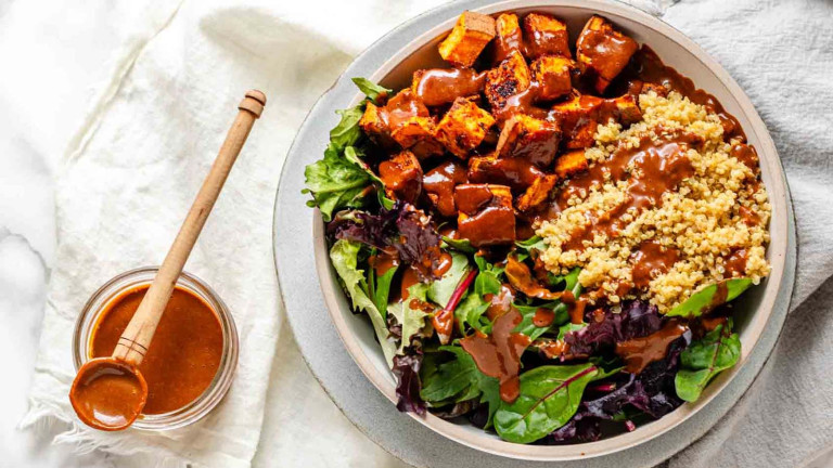 A bowl grains, sweet potatoes and greens, drizzled with peanut sauce.