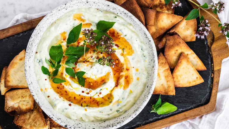 White dip in a bowl garnished with orange sauce and fresh oregano leaves and flowers.