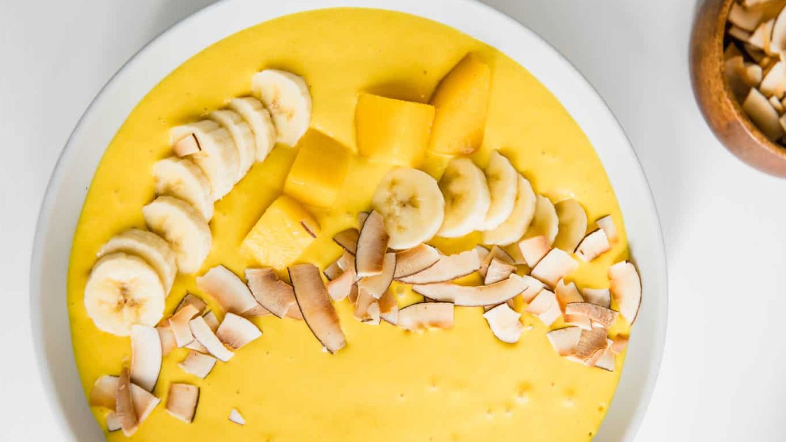 A yellow smoothie bowl topped with banana slices, mango, and coconut.
