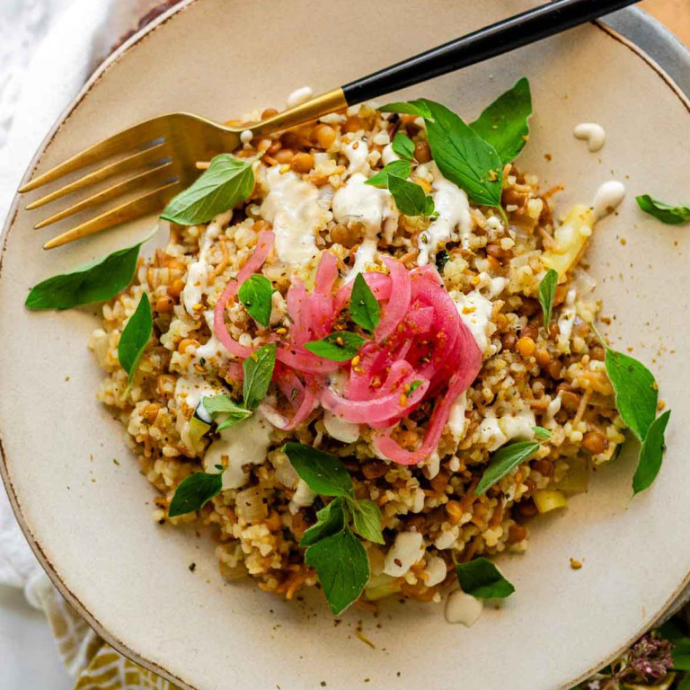 Bulgur and lentil pilaf on a plate with a fork topped with tahini, oregano leaves, and pickled red onions.