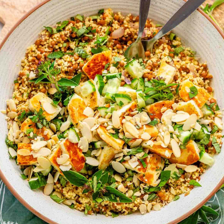 A couscous lentil salad in a bowl topped with grilled halloumi, almond slices, cucumber, and herbs.