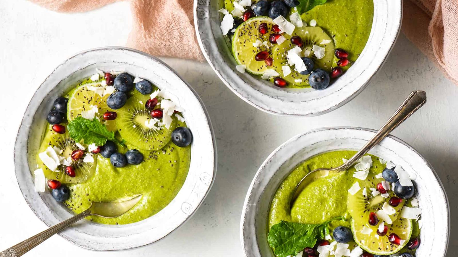 A green smoothie bowl topped with kiwi slices, blueberries, mint, coconut, pomegranate arils, and chia seeds.