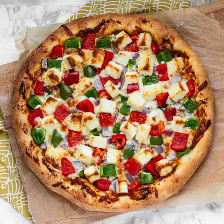 A whole pizza topped with white cubes of paneer and chunks of red and green bell peppers with red onions.