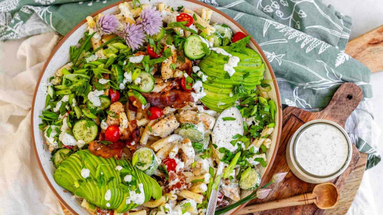 A bowl of pasta salad with chicken, bacon, tomato, avocado, and herbs.