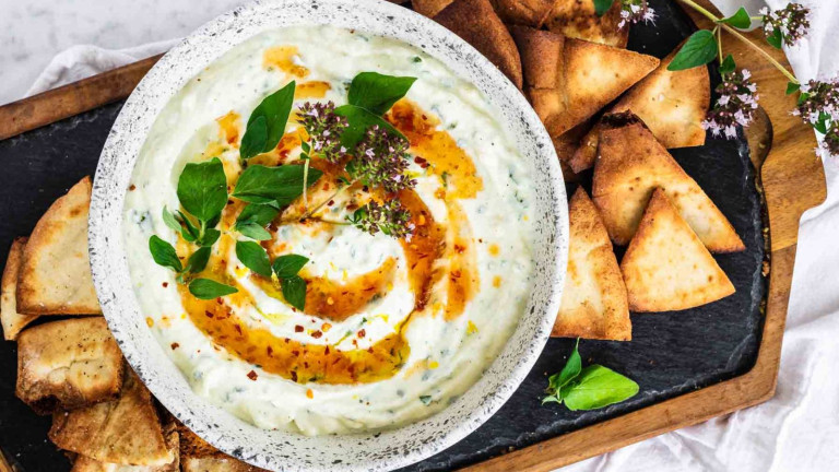 A bowl of white dip topped with an orange swirl of liquid and oregano leaves.