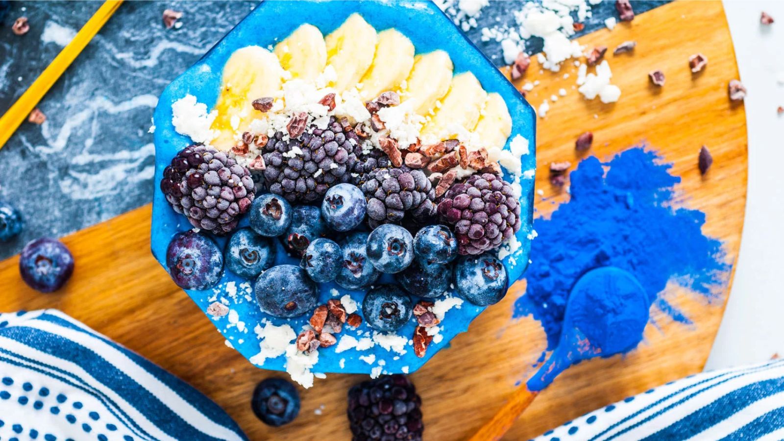 A bright blue smoothie bowl topped with banana slices, nuts, blackberries, and blueberries.