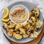 Roasted cauliflower on a plate beside a bowl of dip and lemon wedges.