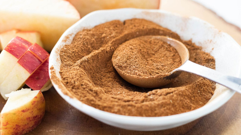 A brown spice mix in a bowl with measuring spoon beside apples.