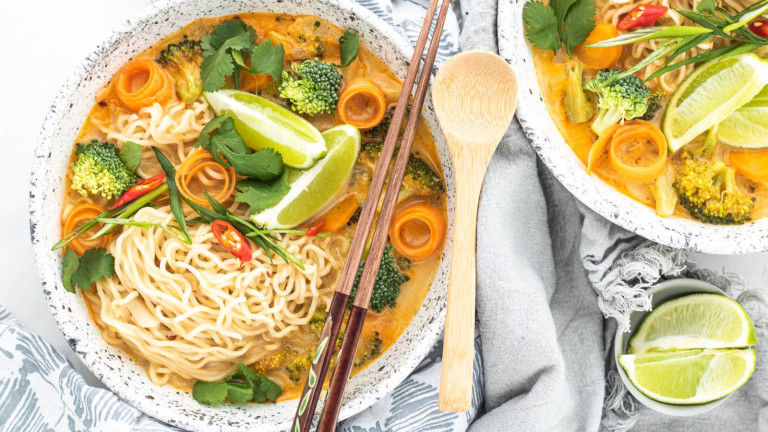 Soup in a bowl with swirled carrots, broccoli, noodles, lime wedges and cilantro.