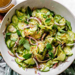 Sliced cucumbers and onions in a bowl with dressing, sesame seeds, and oregano leaves.