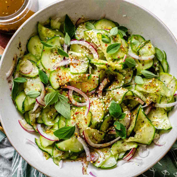 Sliced cucumbers and onions in a bowl with dressing, sesame seeds, and oregano leaves.