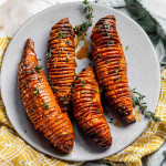 Four sweet potatoes on a plate that have been thinly sliced but not cut all the way through, topped with fresh thyme.