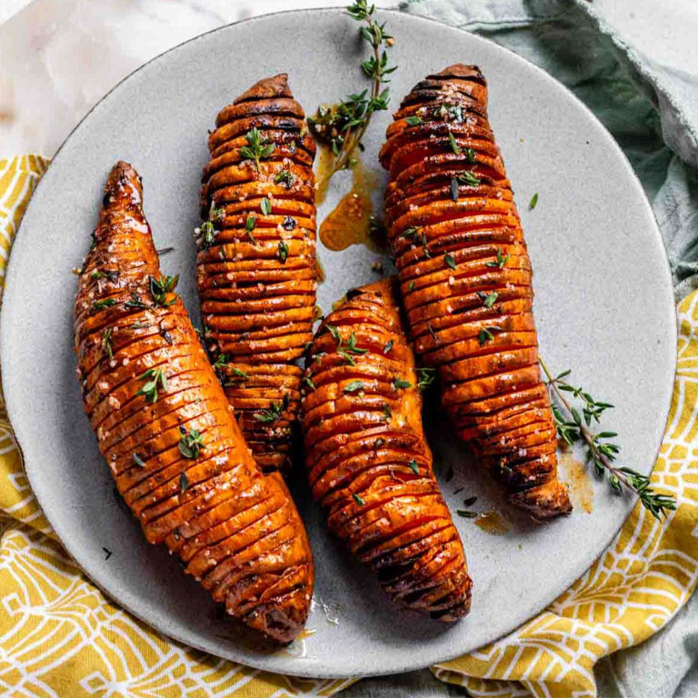 Four sweet potatoes on a plate that have been thinly sliced but not cut all the way through, topped with fresh thyme.