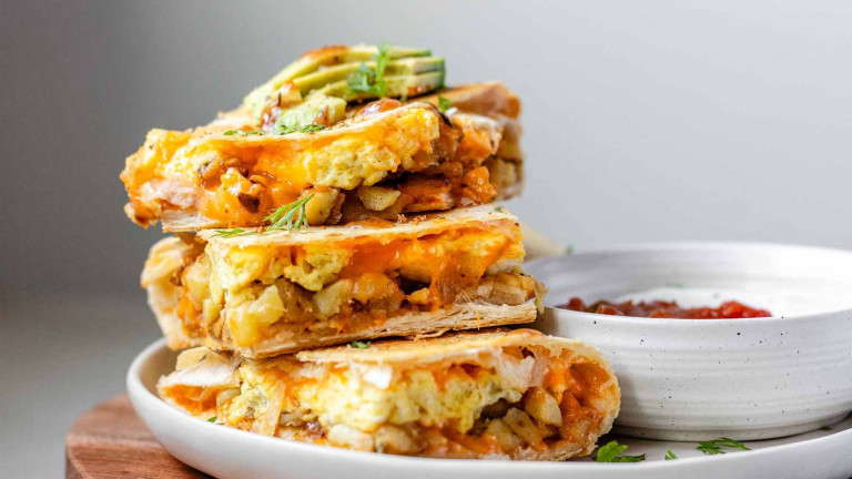 Stacked squares of a tortilla bake topped with avocado slices beside a bowl of salsa and sour cream.