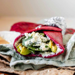 A pink wrap stuffed with avocado slices, spinach leaves, and a white sauce beside a bowl of white sauce.