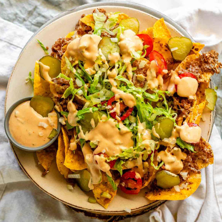 Nachos on a plate topped with lettuce, tomato, beef, onions, pickles, and sauce with a small bowl of sauce.