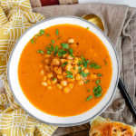 A bowl of orange soup with beans in the middle topped with chopped herbs. An onion bun is beside the bowl.