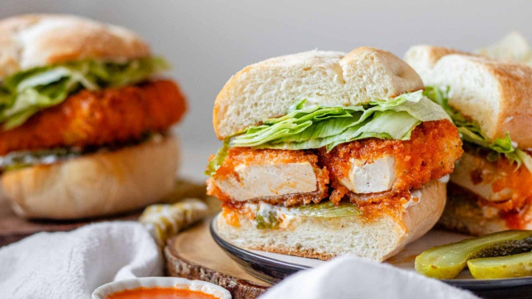 A bun filled with a spicy breaded tofu cutlet, lettuce, sliced pickles, and sauce.