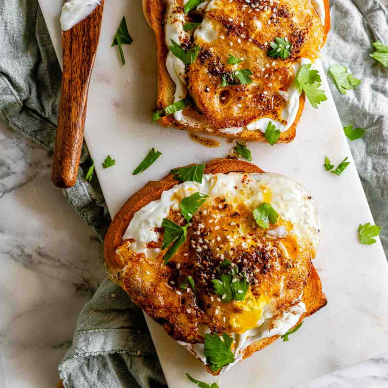 Two slices of toast on a white board topped with white spread, fried eggs, and cilantro.