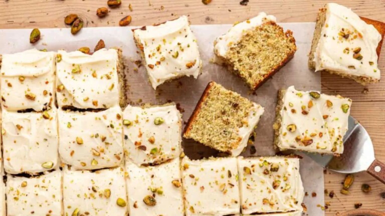 A rectangular cake topped with white icing and chopped pistachios, cut into squares.
